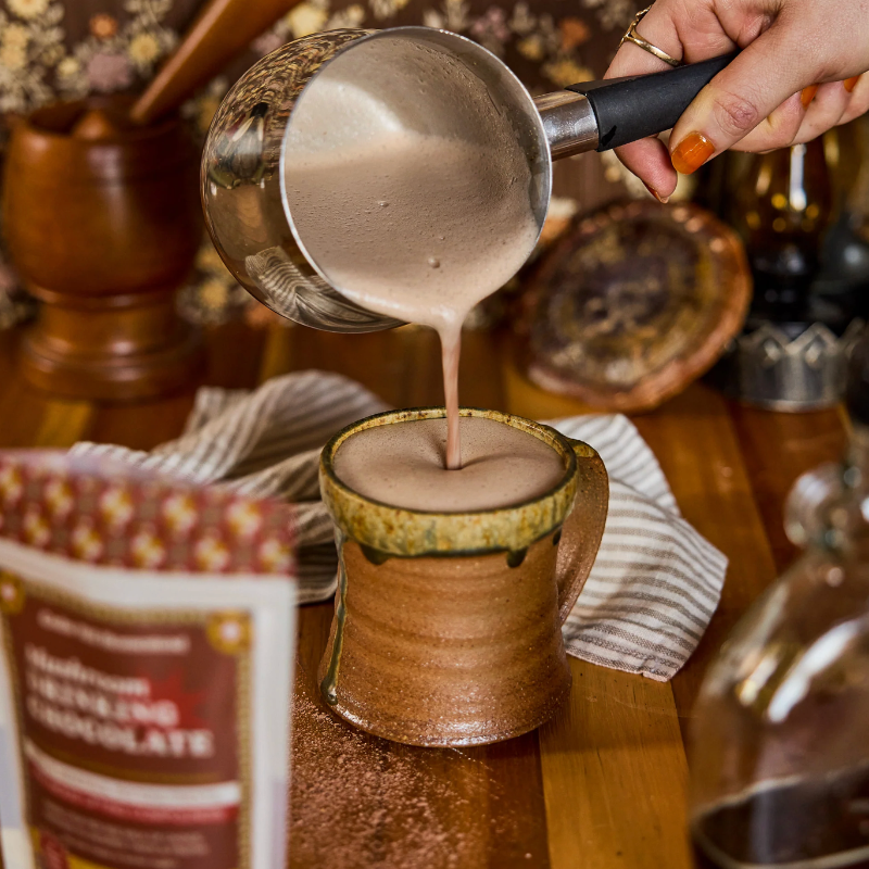Person pouring a creamy liquid into a ceramic mug with a package of chocolate mix in the foreground.